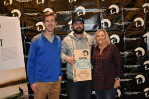 Three people standing in front of a Supreme Boats banner with a plaque.