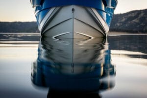 A close-up of the bow of a Supreme S220 boat with blue and white stripes reflecting on calm, still water.