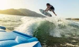 A person is performing an aerial trick on a wakeboard over a wave, with a Supreme Towboats S220 gliding in the background against the sunny coastline and hills.