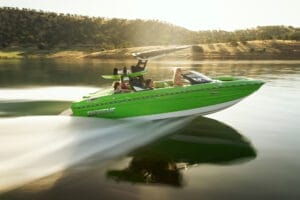 A green Supreme Towboats S220 speedboat with several people onboard travels at high speed on a calm lake, with a hilly landscape in the background on a sunny day.