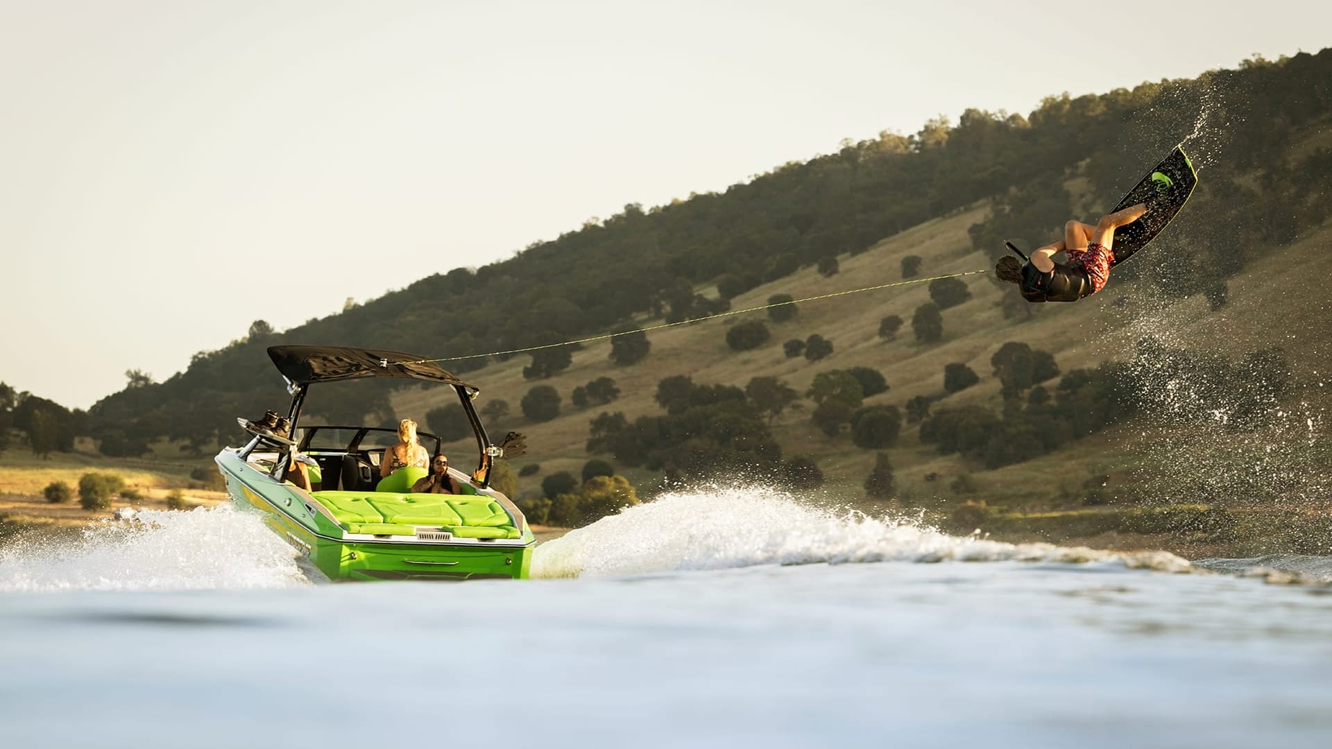 A person is wakeboarding behind a green Supreme Towboats S220 on a lake, with a hilly landscape in the background.