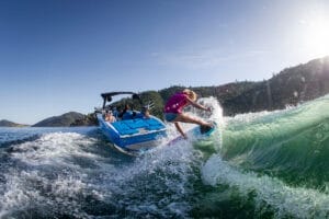 A person in a pink shirt is wakeboarding on a wave created by a blue Supreme S240 boat, with several people on board, in a lake surrounded by hills.