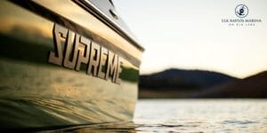 Close-up of a green Centurion boat on a lake with "Supreme" written on its side. The Elk Rapids Marina logo is in the upper right corner of the image. In the background, hills are visible at sunset.