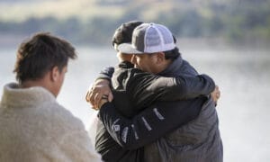 Two men, wearing casual outdoor clothing, share a hug near a body of water with another man standing nearby, embracing the Supreme Life.