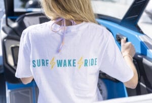 Person in a white T-shirt with "SURF WAKE RIDE" lettering, sitting at the helm of a blue boat, experiencing The Supreme Life, and facing away from the camera.