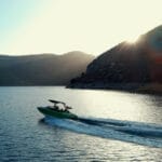 A green speedboat moves quickly across a calm lake surrounded by hills at sunset.