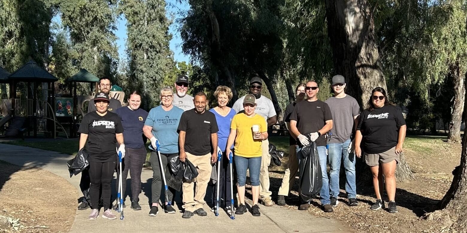 A group of employees stands on a park path, clutching trash bags and tools, poised for a community clean-up. The backdrop reveals trees and a pavilion, as these volunteers embody the spirit of Centurion & Supreme Boats in giving back to their community.