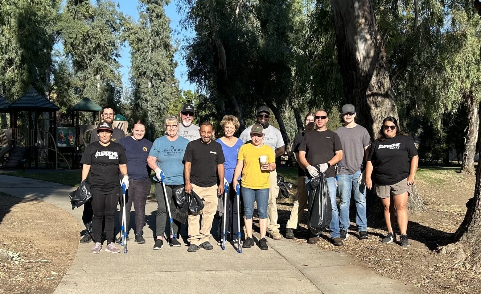 A group of employees stands on a park path, clutching trash bags and tools, poised for a community clean-up. The backdrop reveals trees and a pavilion, as these volunteers embody the spirit of Centurion & Supreme Boats in giving back to their community.