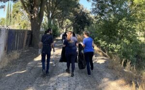 Four people walk on a dirt path lined with trees, carrying trash bags under a clear blue sky, as part of a Community Clean Up effort.