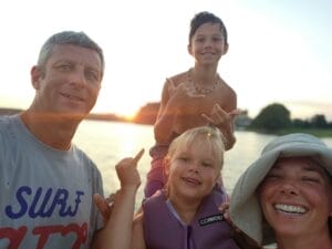 A family of four on a boat at sunset, crafting memories; a man, two children making hand signs, and a woman smiling as they embrace Unger's Supreme Story. Water and trees provide the perfect backdrop for this serene moment.