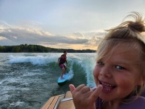 Child making waves surfing behind a boat on a lake, while another child in the foreground smiles and gestures with a hand, capturing unforgettable memories.