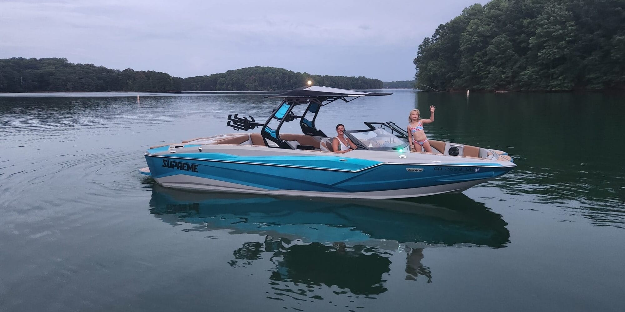 A blue and white boat glides on a calm lake with wooded shores, creating gentle waves; a person stands at the bow, waving as if to capture memories for Unger’s Supreme Story.