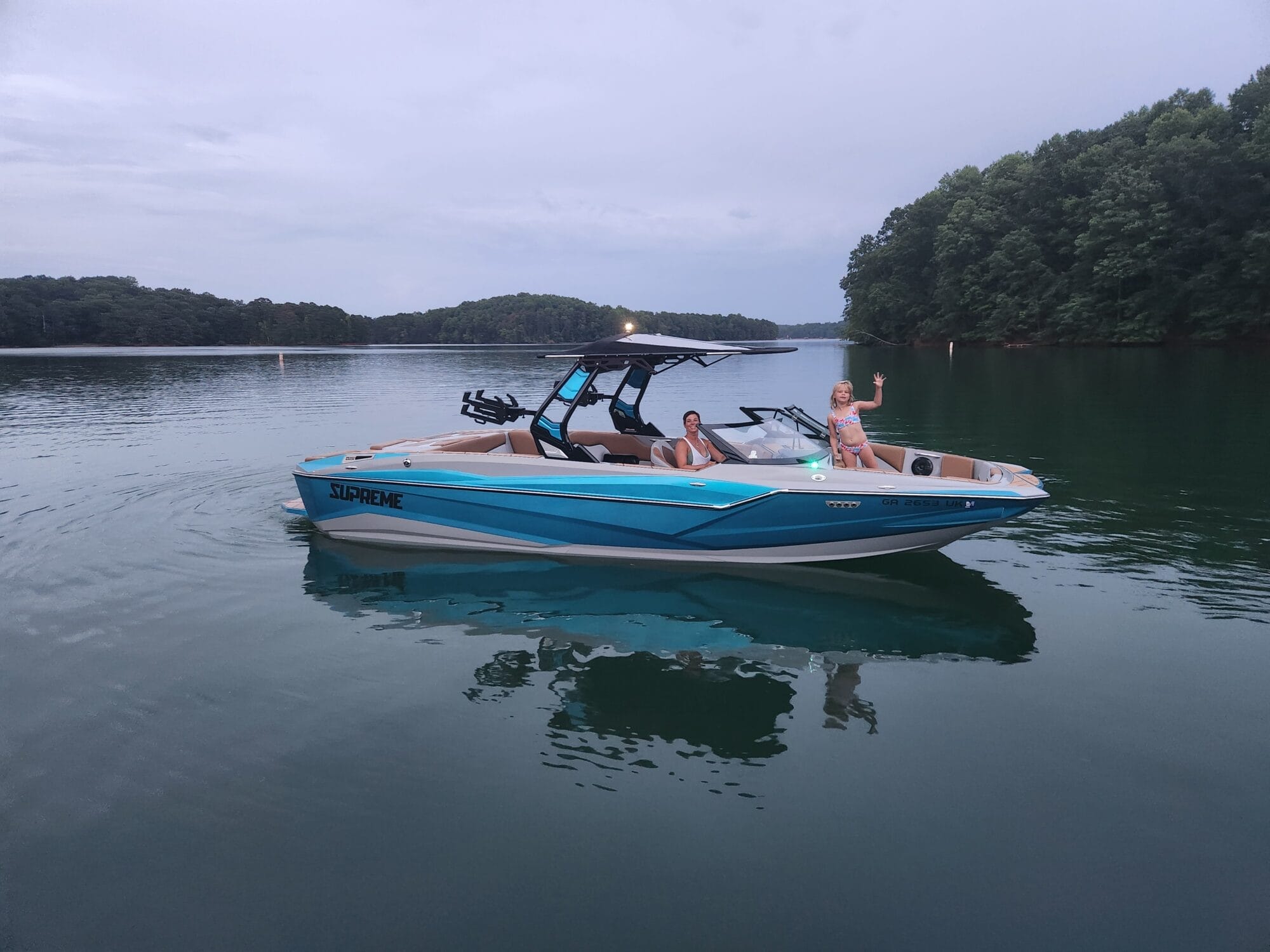 A blue and white boat glides on a calm lake with wooded shores, creating gentle waves; a person stands at the bow, waving as if to capture memories for Unger’s Supreme Story.