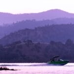 A Centurion green boat speeds across a lake at sunset, with hills silhouetted in the background under a purple sky.