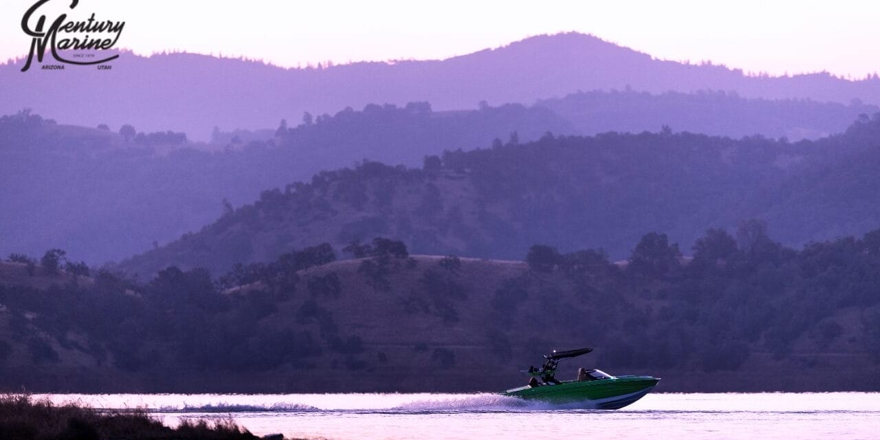 A Centurion green boat speeds across a lake at sunset, with hills silhouetted in the background under a purple sky.