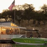 A green boat is docked near a small building adorned with string lights and an American flag, set against a backdrop of trees and a sunset sky—a serene preview to the 2025 Supreme Boat Show extravaganza.