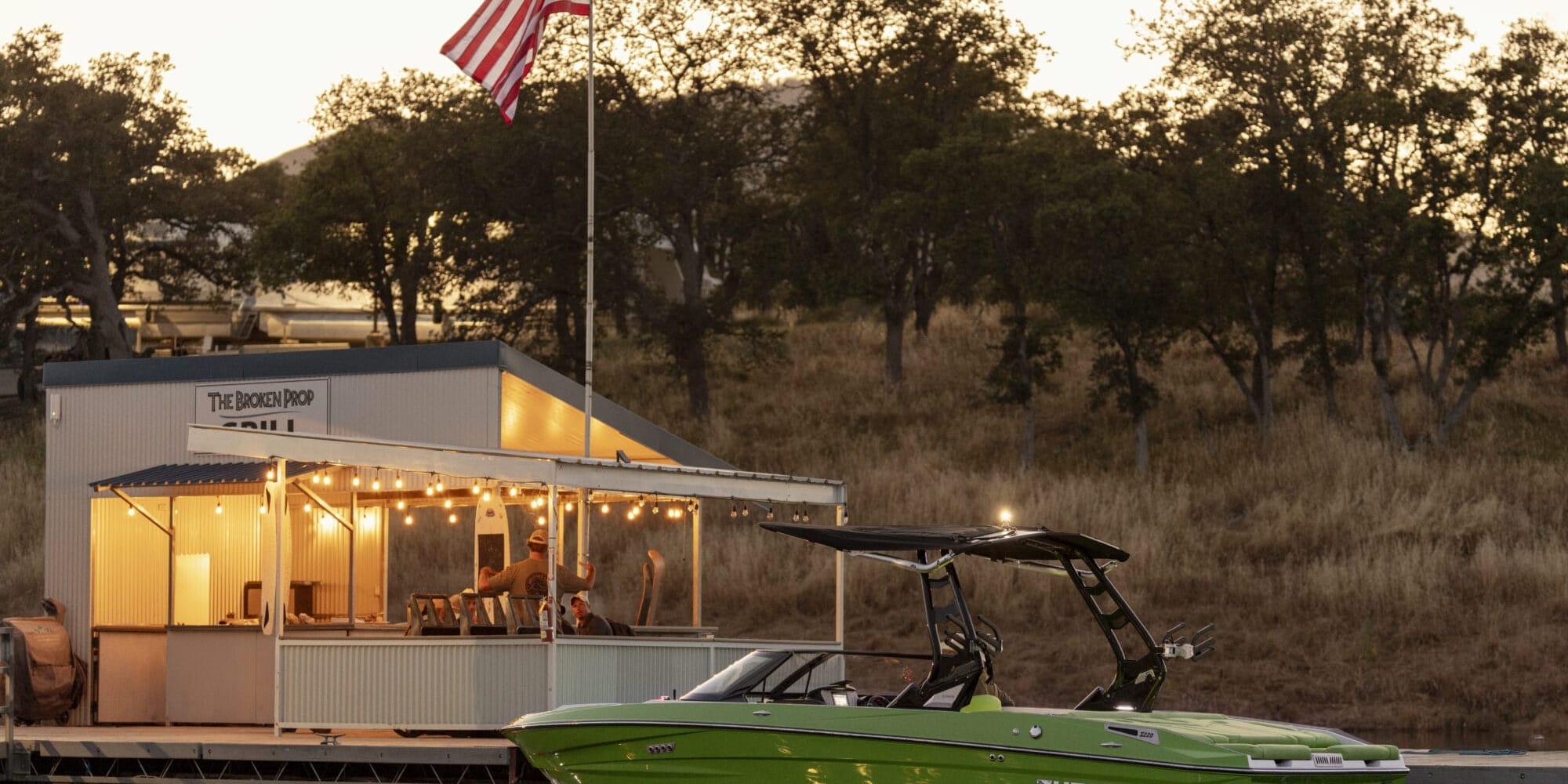 A green boat is docked near a small building adorned with string lights and an American flag, set against a backdrop of trees and a sunset sky—a serene preview to the 2025 Supreme Boat Show extravaganza.