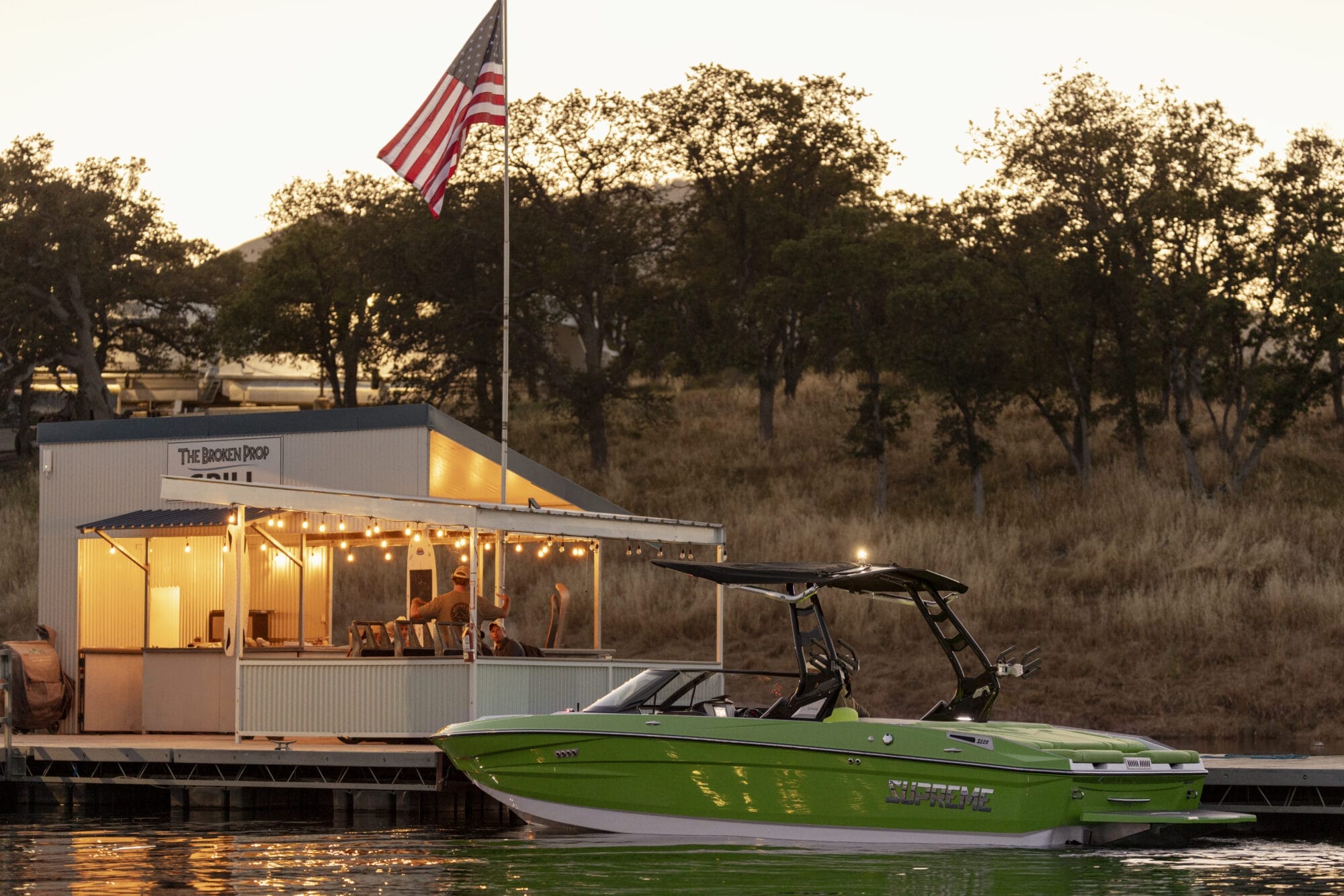A green boat is docked near a small building adorned with string lights and an American flag, set against a backdrop of trees and a sunset sky—a serene preview to the 2025 Supreme Boat Show extravaganza.