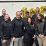 A group of people stands indoors in front of large gold balloon numbers and silver wall panels, all proudly wearing black shirts emblazoned with the Centurion logo, smiling at the camera.