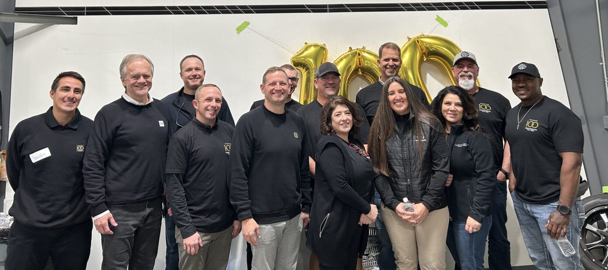 A group of people stands indoors in front of large gold balloon numbers and silver wall panels, all proudly wearing black shirts emblazoned with the Centurion logo, smiling at the camera.