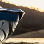 Close-up of a blue and white boat in calm water at sunset, with silhouetted hills in the background and the Oak Hill Marina logo in the corner.