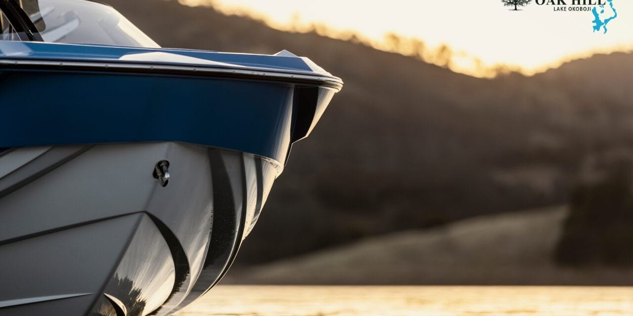 Close-up of a blue and white boat in calm water at sunset, with silhouetted hills in the background and the Oak Hill Marina logo in the corner.