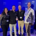 Four people pose in front of a blue backdrop adorned with "WSLA Summit" logos, dressed in casual business attire, as the dim lighting highlights the moment. Among them is the Western Sales Manager, proudly acknowledged for their recent WSIA Leadership Award.